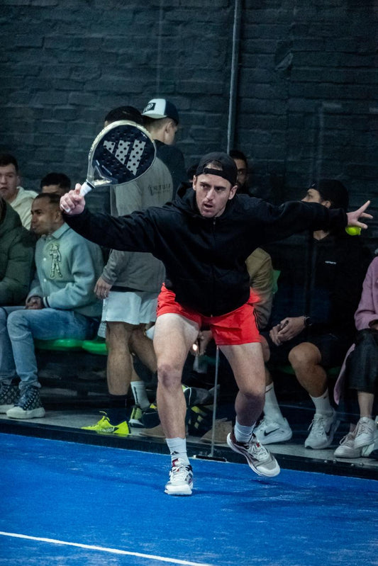 Warren Kuhn hitting a forehand during a competitive Padel match, wearing red shorts and a black hoodie on an indoor court in South Africa.